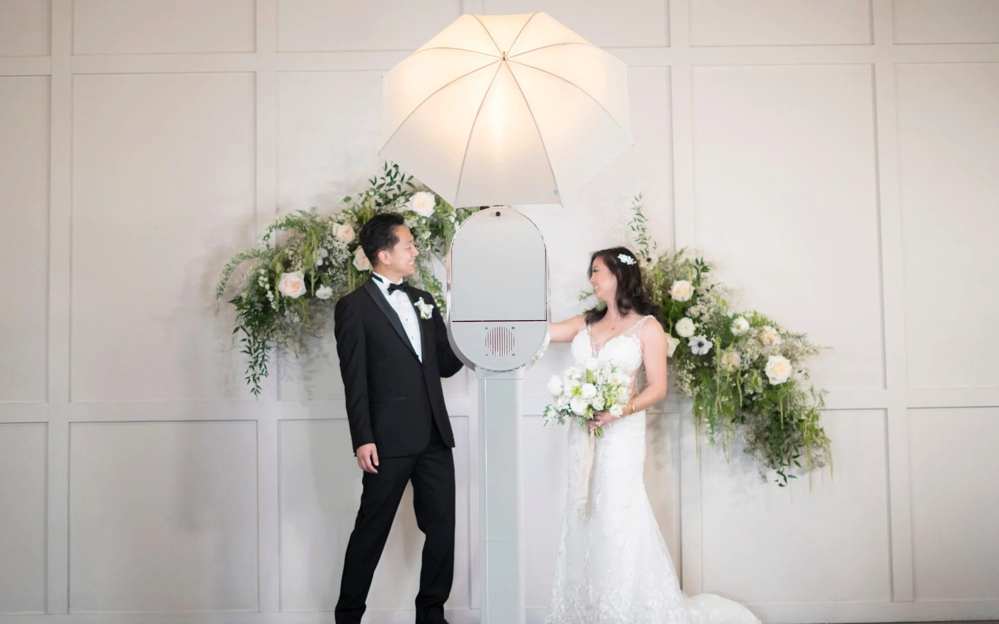 Bride and groom sharing a candid moment inside a stylish wedding photo booth decorated to match a rustic wedding theme, surrounded by guests laughing and holding props.